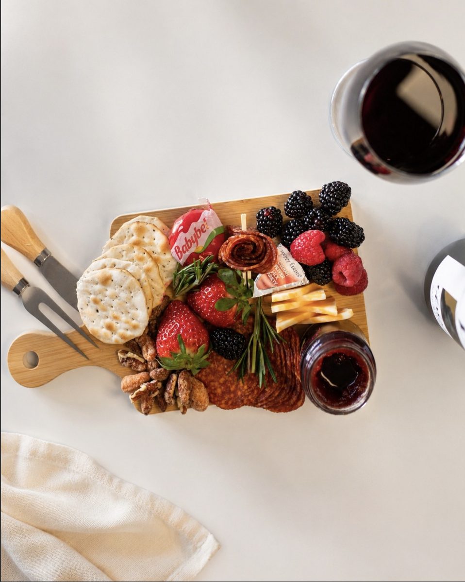 A wooden board of salami, cheese, crackers, strawberries and blackberries beside a glass of red wine.