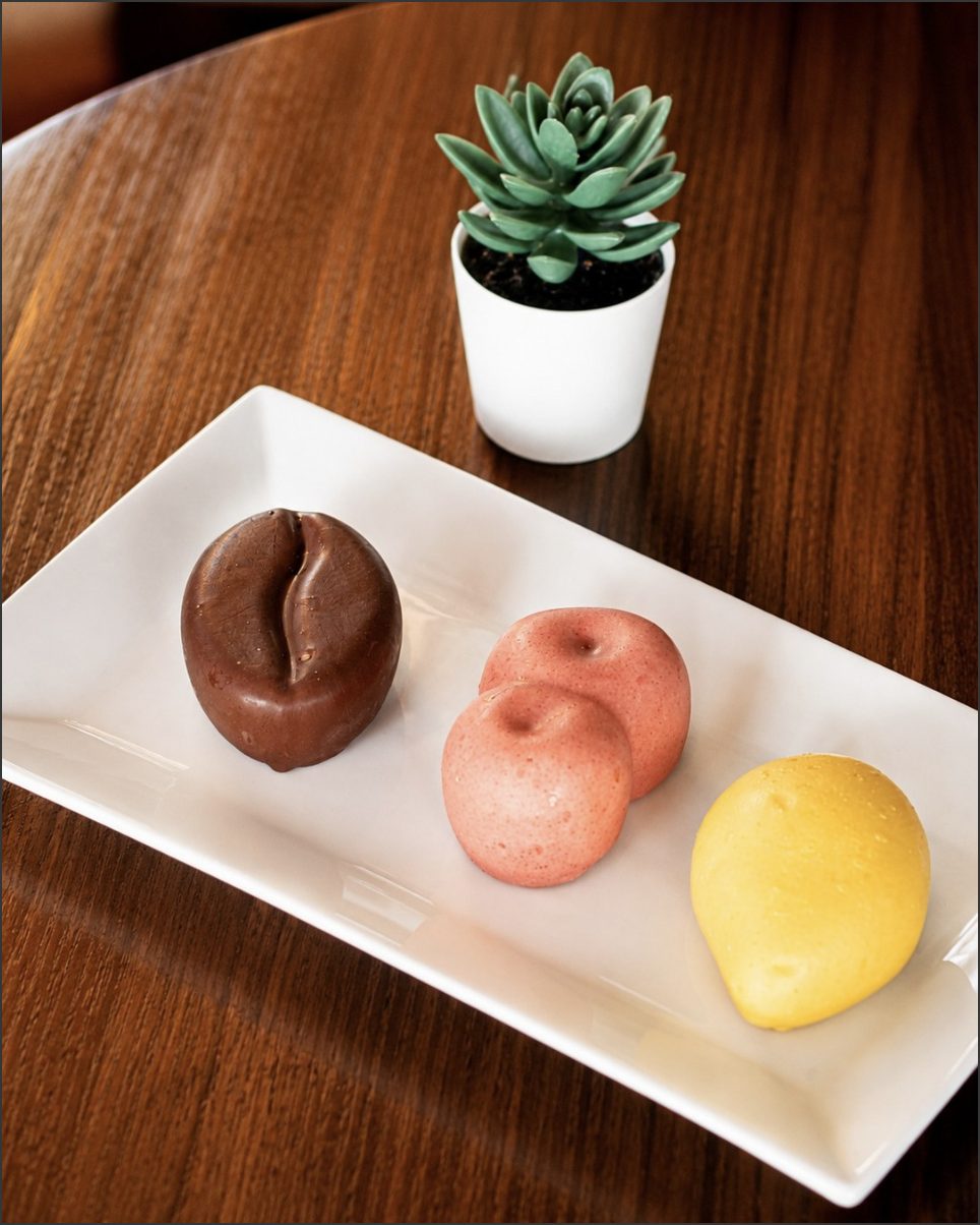 Three glazed mousse desserts — coffee bean, strawberry and mango — on a white plate beside a small succulent.