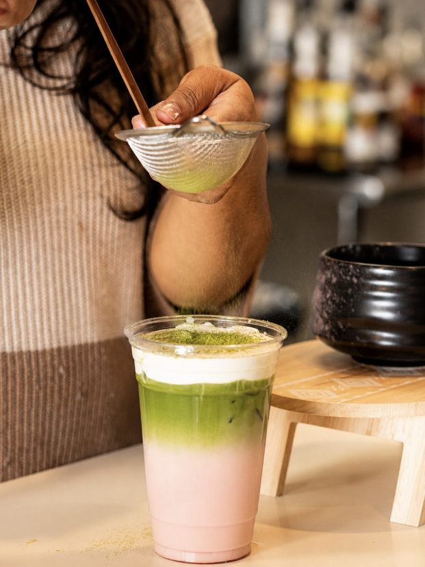 A barista dusting ceremonial matcha through a fine sieve over a layered pink and green strawberry matcha latte.